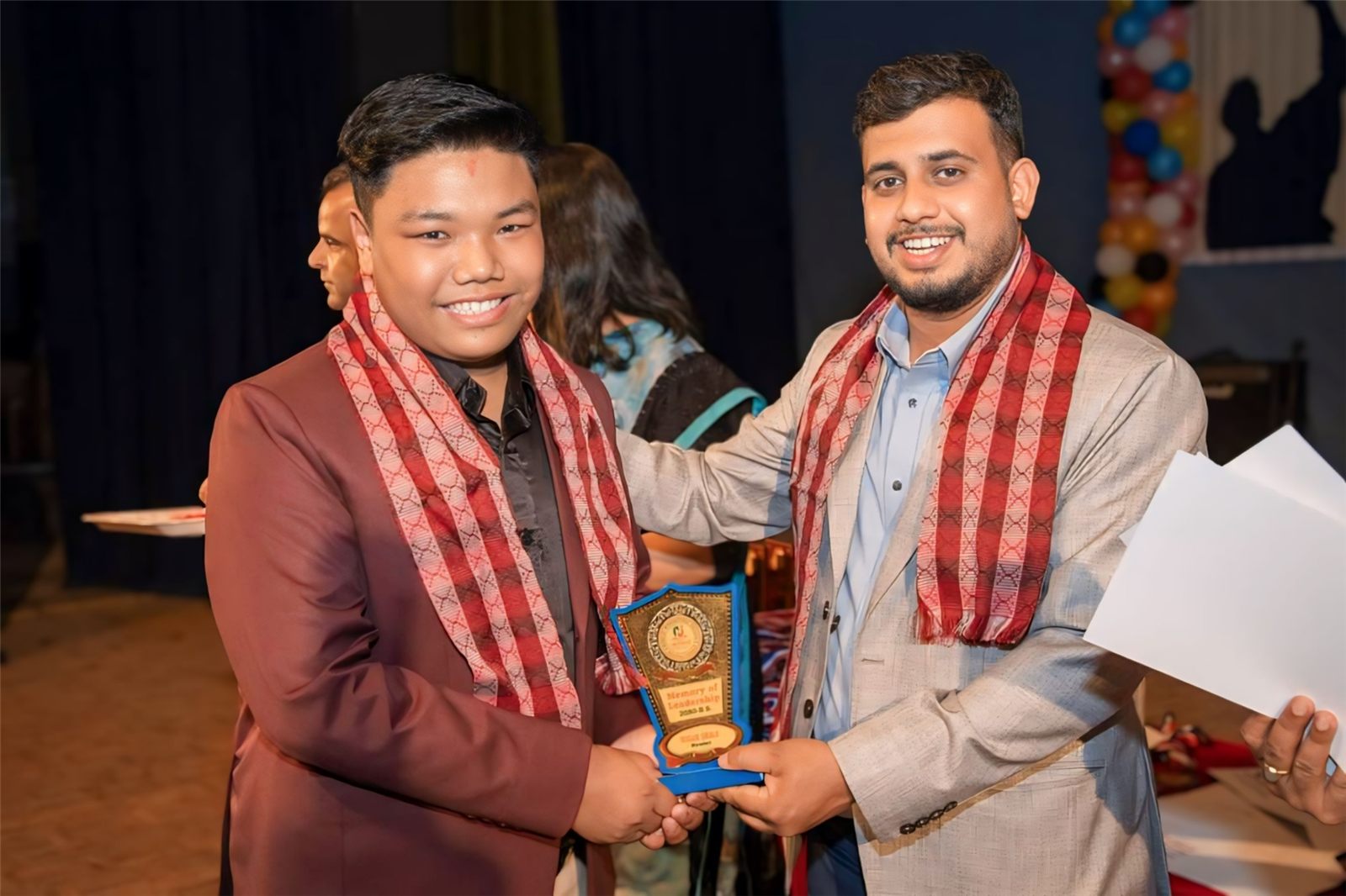 Sugam Ghale receiving a Memory of Leadership trophy on stage, both wearing red patterned scarves
